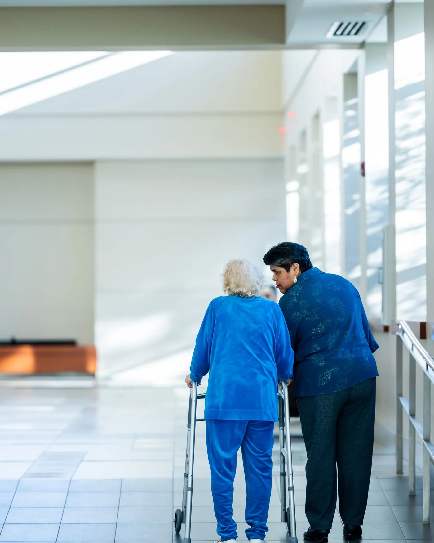 A healthcare aide supporting an elderly passenger using a walker along a bright medical facility corridor
